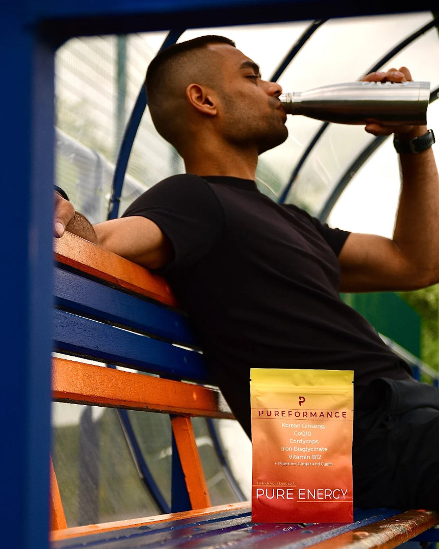 Man drinking from a bottle with a Pureformance Pure energy supplement pouch on a bench.